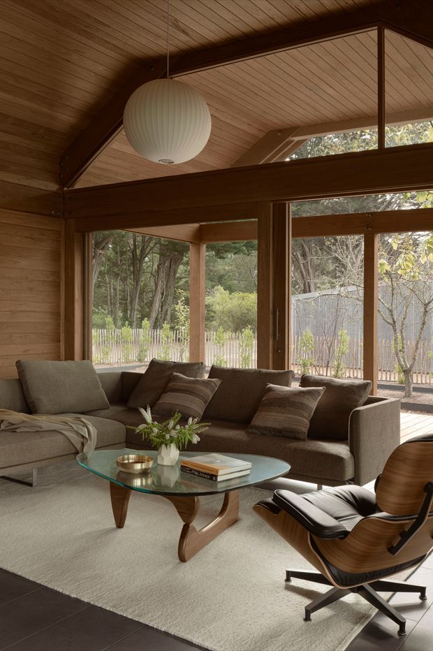 Warm, timber-lined living room with exposed post-and-beam structure, expansive timber-framed glazing, and a high pitched ceiling. The space is furnished with a large taupe sectional sofa, sculptural wooden lounge chair, glass coffee table, neutral rug, and a suspended ribbed pendant light. A leafy landscaped garden is visible through the windows.