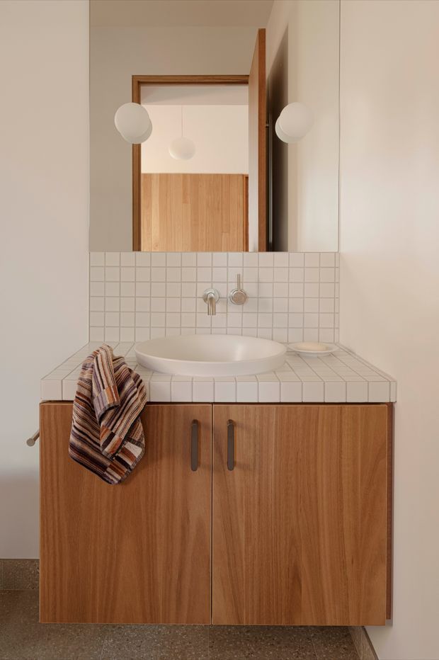 A compact, warmly styled bathroom vanity with a floating timber cabinet, white tiled benchtop and splashback, round vessel basin, wall-mounted brushed metal tapware, and a large mirror. A striped hand towel hangs from the vanity, while the mirror reflects timber joinery and spherical wall lights in the adjacent space.