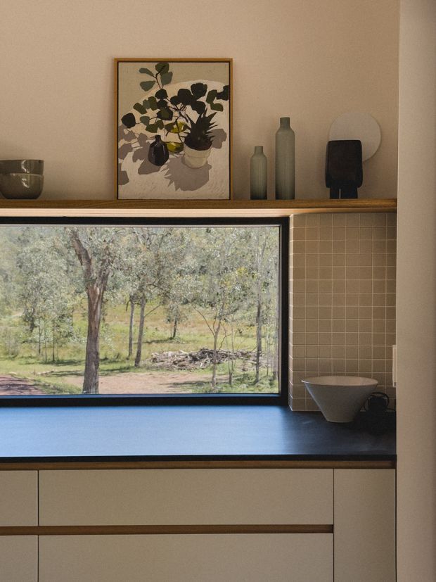 Warm, moody close-up of a contemporary kitchen or kitchenette with flat-panel beige cabinetry, a dark blue benchtop, and a small square beige tiled splashback. A large black-framed window overlooks a wooded natural landscape. Above the window is an open timber shelf styled with a framed botanical artwork, ceramic vessels, and sculptural objects.