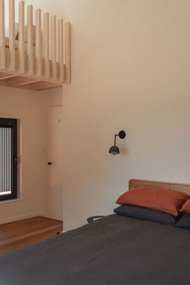 A warm, minimalist bedroom with a dark upholstered bed, rust and charcoal bedding, a simple timber headboard, and a black wall-mounted reading light. A timber staircase or loft railing with vertical slats is visible above the doorway, alongside a dark-framed window and timber flooring. The room has a calm, understated contemporary character with warm neutral walls and natural wood details.