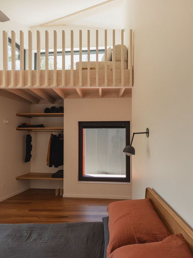 A compact, warm-toned bedroom with a built-in open wardrobe and a timber mezzanine sleeping platform above. Vertical timber slats form the mezzanine balustrade, with exposed timber joists beneath. The room features timber flooring, white plaster walls, a small framed window with translucent ribbed glazing, a simple black wall-mounted reading lamp, and a low timber bed with rust-coloured and charcoal bedding.