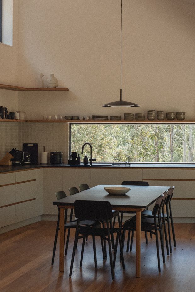 Warm, understated kitchen-dining space with full-height beige walls, timber flooring, pale built-in cabinetry, dark benchtops, open timber shelving, and a long horizontal window overlooking trees. A large rectangular timber dining table with black chairs occupies the foreground, while a slim suspended pendant light hangs over the room. The interior has a restrained mid-century and contemporary character with natural materials and muted earthy tones.