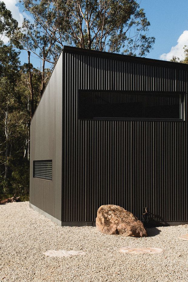 A compact dark-clad building or pavilion viewed from outside, set among eucalyptus trees and a pale gravel landscape. The facade has strong vertical ribbed metal cladding, a long recessed horizontal opening, and a smaller louvred window on the side. A large natural boulder sits against the facade, reinforcing the rugged bush setting. The image has a minimalist, rectilinear architectural character with high contrast between the black exterior and bright gravel and sky.