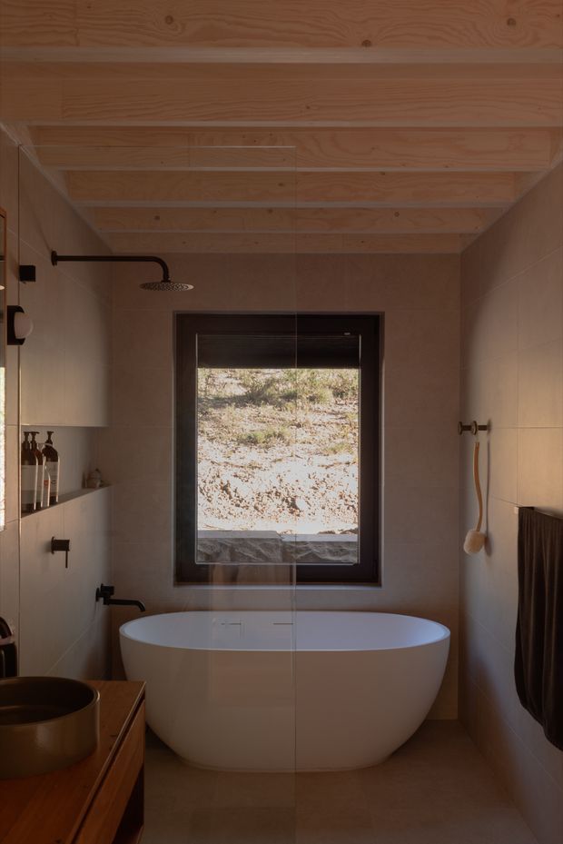A compact, warm-toned bathroom with a freestanding white oval bathtub positioned beneath a dark-framed window. The room has beige tiled walls and floor, black fixtures, a timber vanity with a vessel basin, a wall-mounted shower, and a light timber exposed-beam ceiling. A dark towel hangs on the right wall, while the window looks onto a natural rocky landscape.
