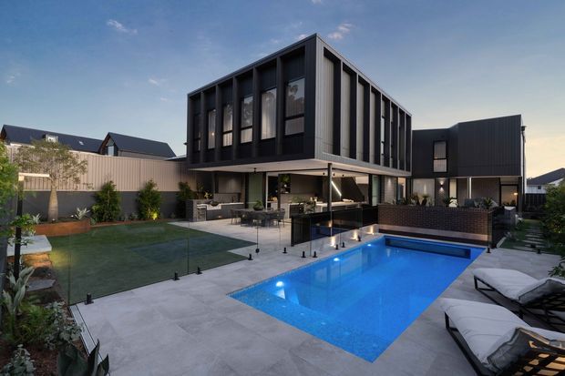 Contemporary multi-storey residential home with a dark, sharply articulated rectangular facade, extensive vertical window framing, and a covered outdoor entertaining area overlooking a brightly lit rectangular swimming pool. The landscaped backyard includes paved pool surrounds, lawn, planting, outdoor seating, and dining furniture. The image is photographed from the exterior at dusk, with the house and pool forming the main composition.
