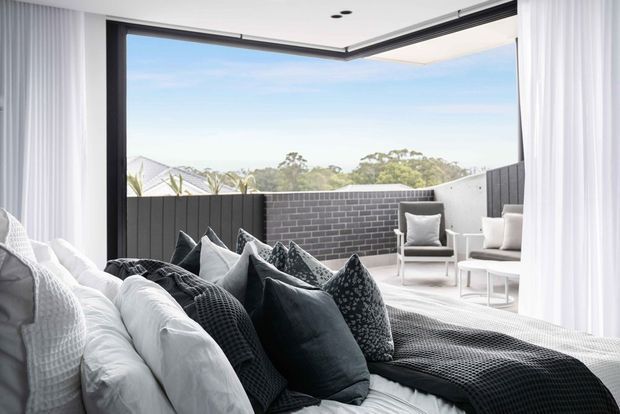 Bright contemporary bedroom with a layered white bed and numerous charcoal and grey cushions, opening through expansive black-framed corner glazing onto a private terrace. Sheer white curtains frame the view, while the terrace has dark grey brick privacy walls and pale outdoor lounge chairs. The room has a calm, monochromatic, airy feel with strong indoor-outdoor connection.