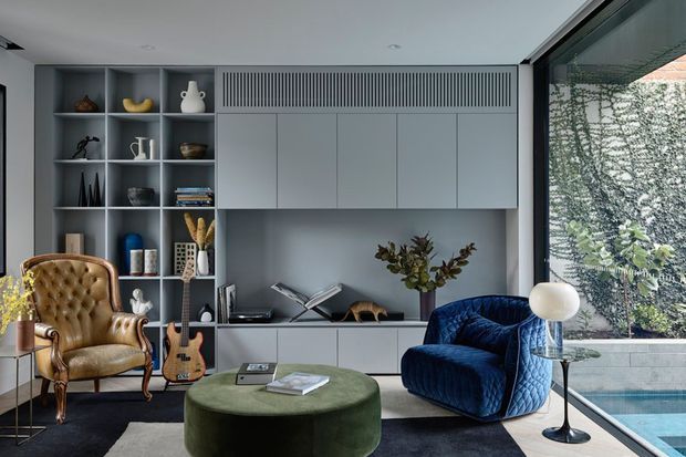 A sophisticated contemporary living room with a built-in grey media and display wall, integrated cabinetry, open shelving, and a large glazed wall overlooking a pool. The room is furnished with a tufted tan leather armchair, deep blue velvet lounge chair, rounded green ottoman, guitar, sculptural decor, and a modern floor lamp. The palette is muted and refined, with strong indoor-outdoor connection and abundant natural light.