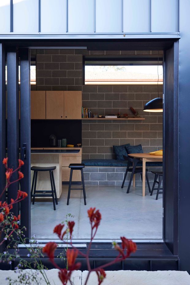 A compact contemporary open-plan living and dining space viewed through a large dark-framed glazed opening. The room features exposed dark grey concrete block walls, polished concrete flooring, light timber kitchen cabinetry, a built-in blue upholstered bench, a timber dining table, and black pendant lighting. A high horizontal window brings in daylight, while the foreground includes blurred red flowering plants outside.