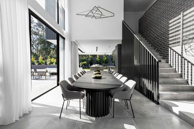 A bright, high-ceilinged contemporary dining area with a large oval dining table and upholstered chairs beside full-height glazing. White curtains, pale stone-look flooring, a dark vertically ribbed table base, and a black brick feature wall with a staircase create a refined monochromatic interior. The glazing opens toward an outdoor terrace and landscaped courtyard, while a geometric pendant light hangs above the table.