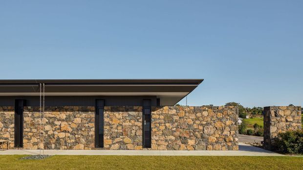 Low, single-storey contemporary building exterior with a broad flat projecting roof and walls constructed from irregular natural stone masonry. Dark vertical structural elements and a narrow concrete walkway contrast with the warm, variegated stone facade. The image focuses on the building envelope and roofline, with landscaped grounds and distant terrain in the background.