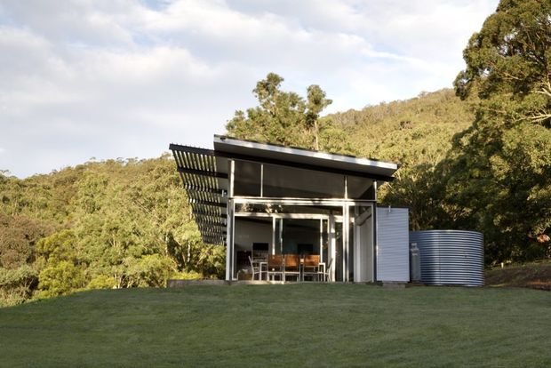 A compact modern pavilion set within a grassy clearing and surrounded by dense wooded hills. The building has a flat, slightly projecting roof, extensive floor-to-ceiling glazing, a deep slatted sun-shading screen along one side, and corrugated metal cladding. A large cylindrical corrugated water tank sits beside the structure.
