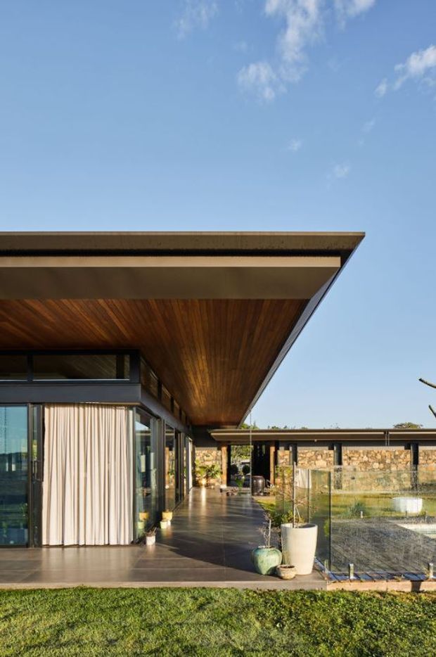 Exterior view of a low-slung contemporary house centred on a broad cantilevered roof canopy with a warm timber-lined soffit. Full-height glazing, dark slender window frames, stone-clad walls, covered outdoor circulation, and a reflective water feature surround a paved courtyard-like terrace. The scene is sunlit beneath a clear blue sky, with lawn in the foreground.