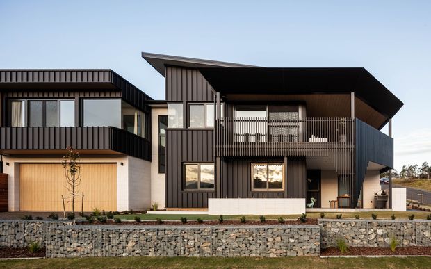 Contemporary multi-level residential house viewed from the front, featuring dark vertical cladding, angular roof forms, projecting balconies with slim metal balustrades, large windows, and an integrated garage. The home sits above a landscaped gabion retaining wall and paved forecourt.
