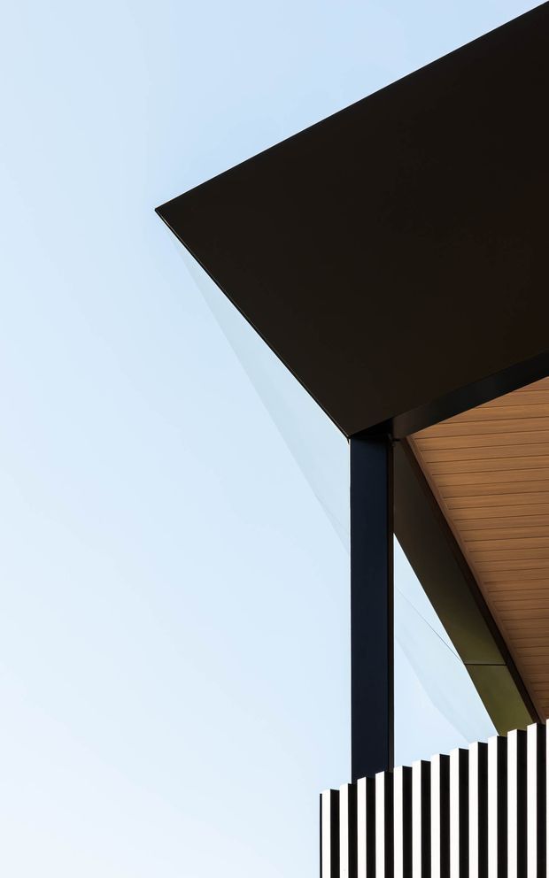 Minimal architectural exterior detail showing a sharply cantilevered dark roof plane supported by a slender black post, with warm timber soffit lining and a vertical black-and-white slatted fence against a pale blue sky.