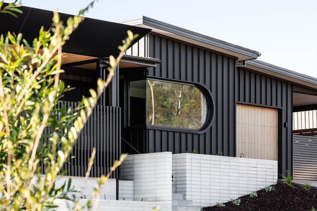Exterior view of a contemporary single-storey residence with dark vertical cladding, a low-pitched roof, distinctive rounded-corner window, covered entry, and pale brick boundary walls. Landscaping partially frames the foreground.