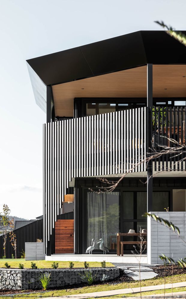 Exterior architectural photograph of a contemporary multi-level house featuring a dramatic black folded roof, warm timber soffit, black-framed glazing, vertical black-and-white screening, timber-clad details, and gabion retaining walls with landscaped planting.