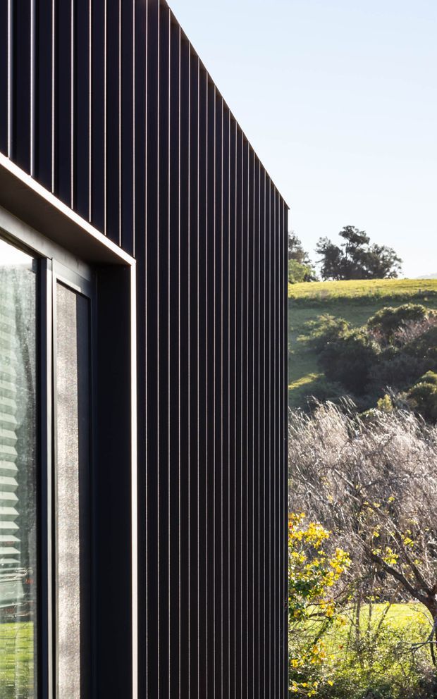 Close-up exterior architectural view of a contemporary building with a tall, dark vertically ribbed facade and a deeply recessed glazed opening. The facade forms a sharp geometric edge against a bright sky and landscaped rolling terrain, with strong sunlight highlighting the vertical cladding lines.