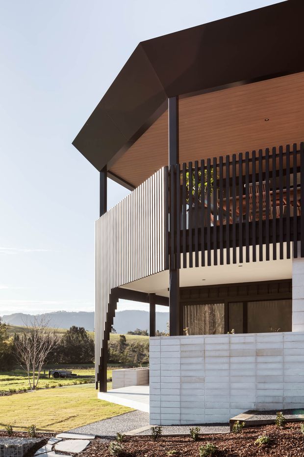 Exterior architectural photograph of a contemporary multi-level house with a dramatic projecting roof, warm timber-lined soffits, dark vertical slatted screens and slender dark structural posts. A pale masonry wall forms the lower level, with a covered terrace or balcony overlooking a landscaped rural setting and distant hills.