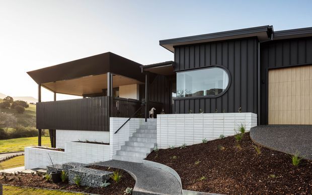 Modern two-level residential house on a sloped site, featuring dark vertical cladding, a dramatic curved picture window, a covered upper terrace with slender posts and privacy screening, pale masonry retaining walls, and a curving paved entry path. The architecture has strong horizontal and vertical geometric forms with a restrained black, white, and natural landscape palette.