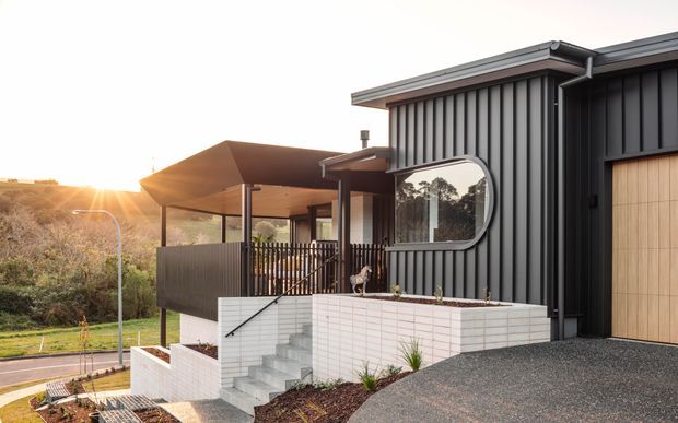 Exterior view of a contemporary dark-clad house on a sloping site, featuring a covered entry terrace, vertical ribbed metal cladding, a large rounded-corner window, pale brick planter walls, and an attached timber garage door. The landscaped setting and low sunset light emphasise the modern residential design.