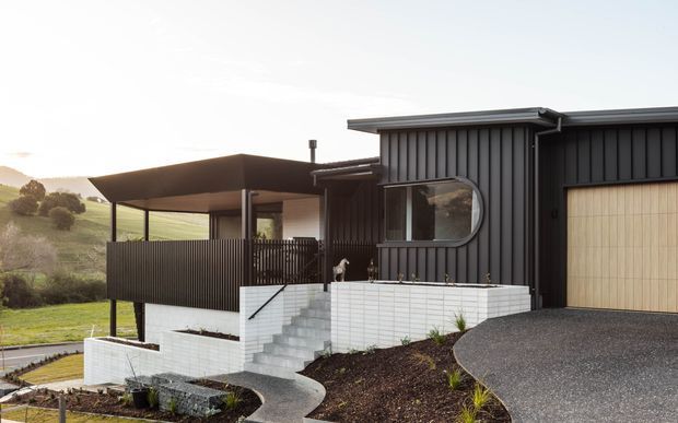 Contemporary dark-clad house exterior on a gently sloping rural site, featuring a low-pitched roof, vertical ribbed cladding, a timber garage door, a covered outdoor terrace with screened balustrade, and white brick retaining walls with concrete steps and curved paths.
