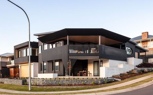Contemporary multi-level residence viewed from the street, featuring dark vertical cladding, expansive covered balconies, strong horizontal roof planes, pale masonry walls, and landscaped front terraces on a sloping suburban site.