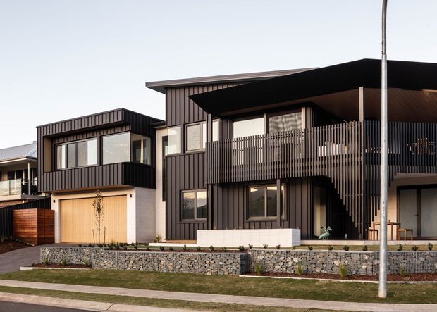 Street-facing view of a contemporary multi-storey residential building with dark vertical cladding, projecting flat roof forms, large windows, a screened upper-level balcony, garage, and stone retaining walls.