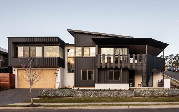 Front exterior view of a contemporary multi-level residence with dark vertical cladding, strong angular roof forms, projecting upper-level balcony, large glazed openings, and a stone retaining wall along the street frontage.