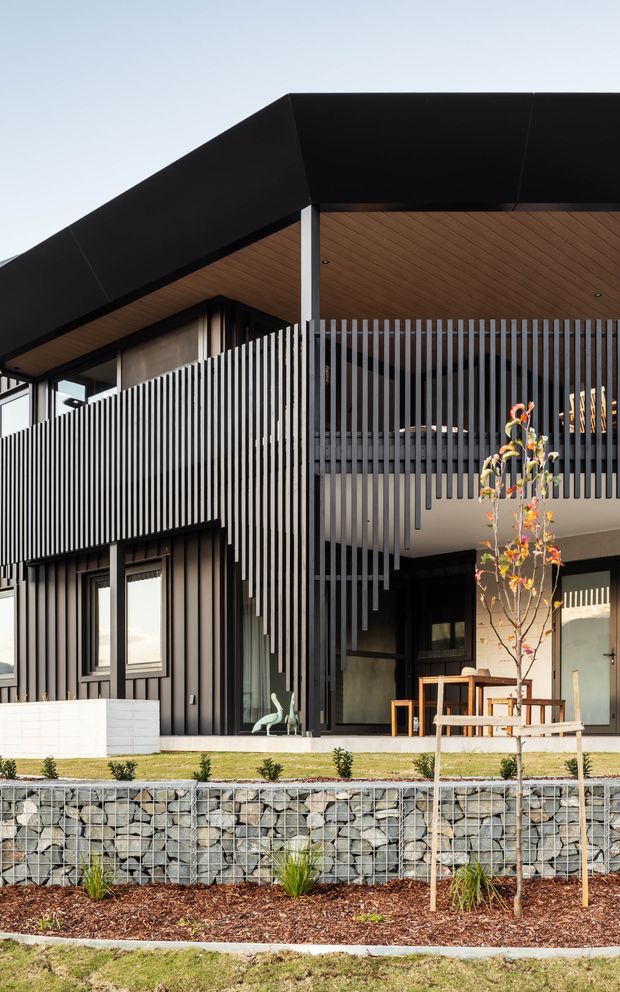 Front exterior view of a contemporary two-storey house with a dramatic black angular roof, timber-lined soffits, dark vertical screening and cladding, extensive glazing, and a recessed sheltered entry or outdoor living area. The property sits above a gabion retaining wall with landscaped planting and a young tree.