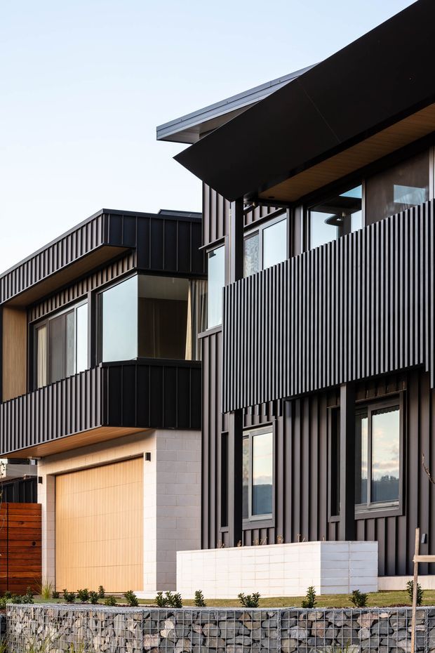 Exterior view of a contemporary multi-storey residential building with strong geometric forms, dark vertical ribbed cladding, projecting black roof planes, extensive glazing, timber soffits, balconies, and pale masonry elements. A gabion retaining wall and landscaped planting line the foreground.