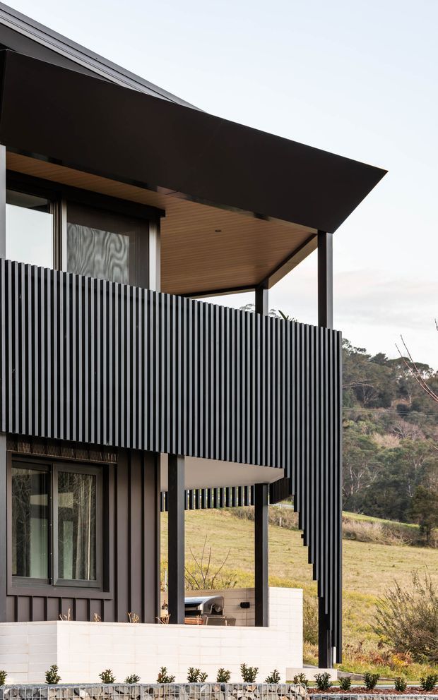 Exterior architectural photograph of a contemporary multi-level house on a sloping rural site. The facade combines dark vertical screening and cladding with large glazed openings, slender dark structural posts, a dramatic projecting flat roof, and a warm timber-lined soffit. A low white masonry wall and landscaped planting sit at the base, with open grassy terrain and trees beyond.