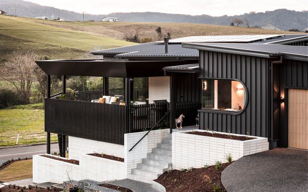 Exterior view of a contemporary multi-level home on a sloping rural site, featuring dark vertical cladding, light brickwork, a corrugated metal roof, large glazed openings, a covered outdoor terrace, and landscaped raised planters. Rolling hills and open countryside are visible behind the property.