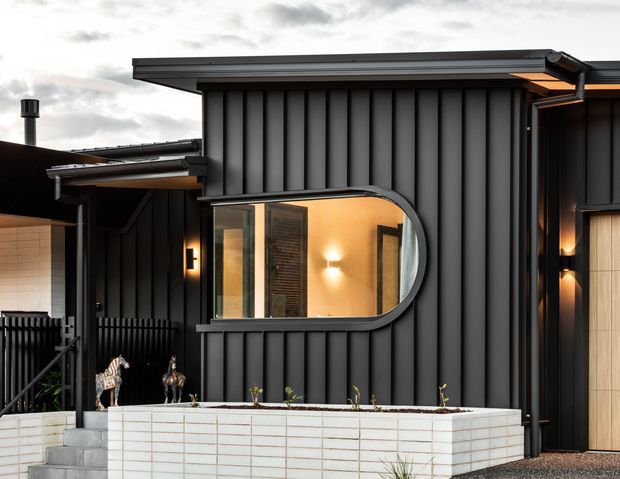 Exterior view of a contemporary dark-clad building with vertical ribbed metal wall panels, a distinctive rounded rectangular window, warm interior lighting, and a white tiled raised planter or wall in the foreground. The composition highlights the facade and its contrasting geometric forms.