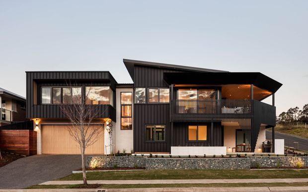 A contemporary multi-level residential house photographed from the street at dusk. The facade combines dark vertical cladding, expansive glazing, a projecting upper balcony, and a sharply angled roof canopy. A timber-toned garage door, low stone retaining wall, landscaped verge, and warm interior lighting add contrast to the dark exterior.