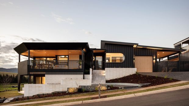 Contemporary multi-level residential house viewed from the street at dusk, featuring dark vertical cladding, a sharply angled roof and covered upper balcony, warm interior lighting, pale masonry retaining walls, a recessed garage, and landscaped sloping frontage. A rural or semi-rural landscape is visible beyond the property.