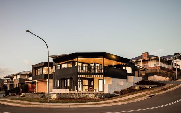 Street-level exterior view of a contemporary multi-storey residential house on a sloped suburban site. The home features dark horizontal cladding, extensive floor-to-ceiling glazing, projecting box forms, open balconies, warm interior lighting, and a mixed flat and low-pitched roof composition. Adjacent modern residences are visible along the street.