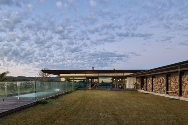 A low-slung contemporary residential property viewed from a broad landscaped lawn at dusk. The building has strong horizontal rooflines, extensive floor-to-ceiling glazing, warm interior lighting, and a stone-clad wall section with timber-toned detailing. A glass balustrade borders a paved terrace or poolside area on the left, with distant hills visible beyond.