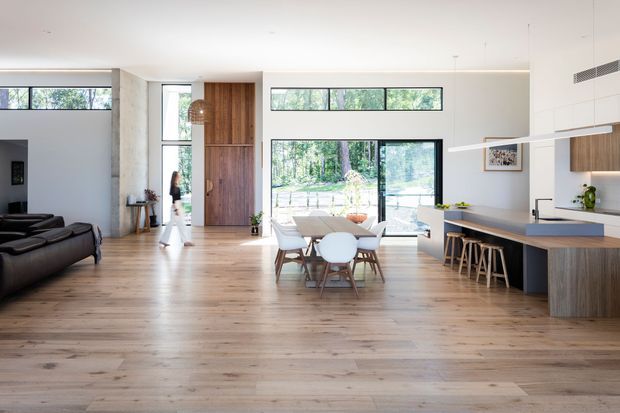 Bright open-plan contemporary home interior combining a kitchen, dining area and living room. The space features warm timber flooring, white walls and cabinetry, a large dark kitchen island with timber detailing, integrated appliances, extensive black-framed glazing, and a tall timber entry door. A dining table sits between the kitchen and living area, with views to a wooded outdoor setting.