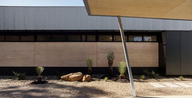 Low, modern building exterior with a strong horizontal composition, dark corrugated metal roof, a band of narrow clerestory windows, large warm-toned wall panels, and a projecting timber-toned canopy supported by a slim angled post. A gravel xeriscape garden with rocks and drought-tolerant plants fronts the facade, alongside pale stepping pavers.