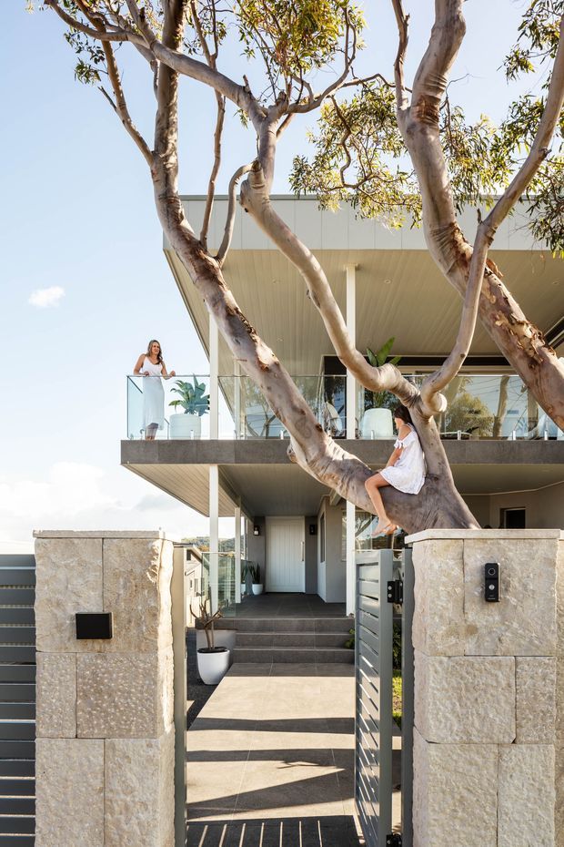 A contemporary multi-level residential facade viewed from the front entrance, framed by mature trees. The home features a broad projecting roof, glass balcony balustrades, expansive glazing, dark tiled entry steps, a metal pedestrian gate, and substantial pale stone masonry pillars and walls. Two people are visible on the balcony and in the tree, adding scale.