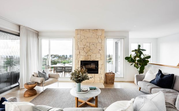 Bright, airy contemporary living room with expansive glazing, a central rough natural-stone fireplace surround, pale walls, light furnishings, a neutral area rug, and views to landscaped surroundings. The room includes a large sectional sofa, armchair, round coffee table, indoor plant, and a secondary seating/dining area near the windows.