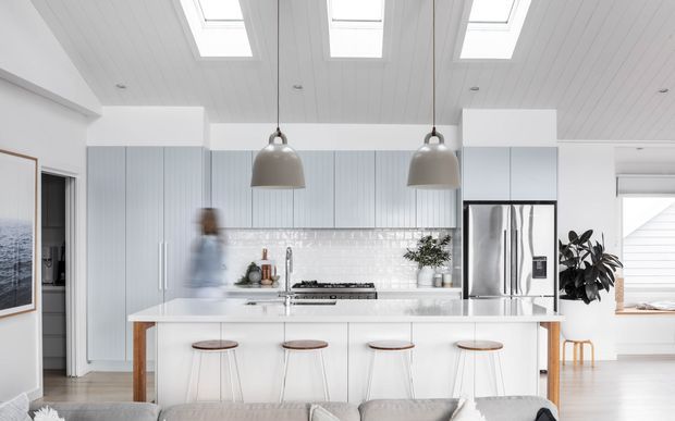 Bright, open-plan contemporary kitchen with a large white island, four stools, pale vertical-panel cabinetry, white tiled splashback, stainless-steel appliances, and twin pendant lights. White timber-look ceiling panels with multiple skylights add natural light, while warm timber accents and indoor plants soften the clean palette.