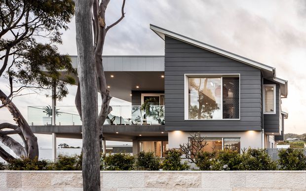 Contemporary two-storey residential house viewed from the street, featuring dark horizontal cladding, a dramatic asymmetrical sloping roof, large windows, glass balcony balustrades, recessed exterior lighting, and a pale masonry boundary wall. Mature trees and landscaped planting frame the facade.