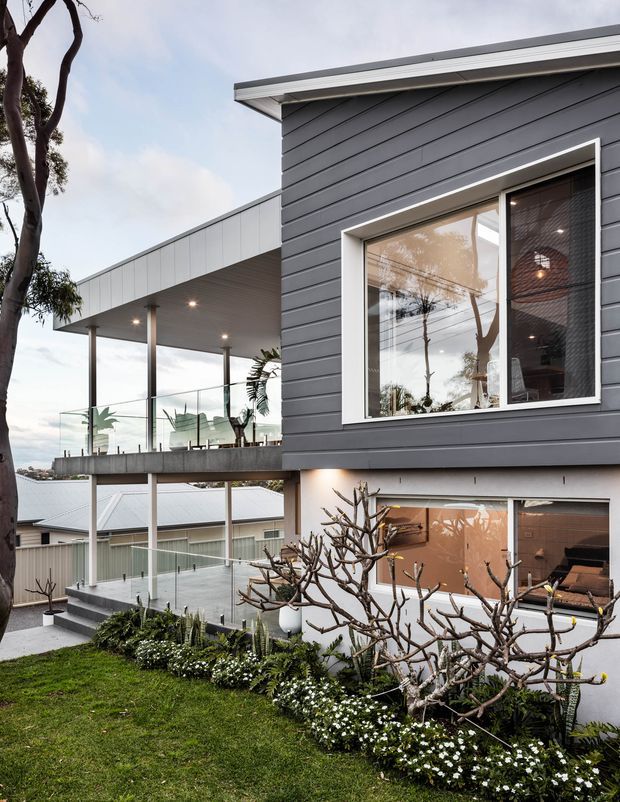 A contemporary multi-level residential exterior on a sloping site, featuring dark grey horizontal cladding, white trim, a broad flat-roofed balcony or terrace with clear glass balustrades, and extensive glazed openings. Landscaped gardens and neighbouring rooftops are visible around the elevated structure.