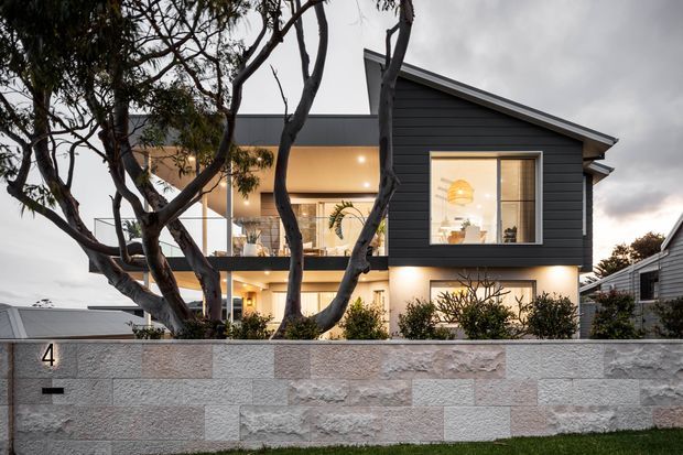 Exterior view of a contemporary two-storey residence at dusk, partially screened by mature trees and a low textured masonry boundary wall. The house combines dark horizontal cladding, expansive glazed openings, warm interior lighting, a projecting upper-level balcony, and a sharply sloping single gable roof.