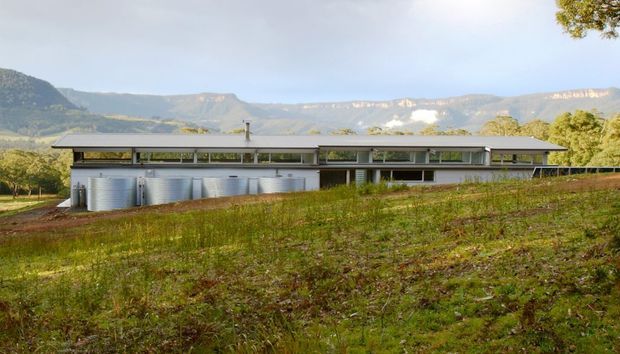 A long, low-slung single-storey building set in a cultivated rural landscape, with a shallow-pitched roof, extensive horizontal glazing, pale walls, and several large cylindrical metal tanks along the side. Dramatic mountain escarpments form the distant backdrop.