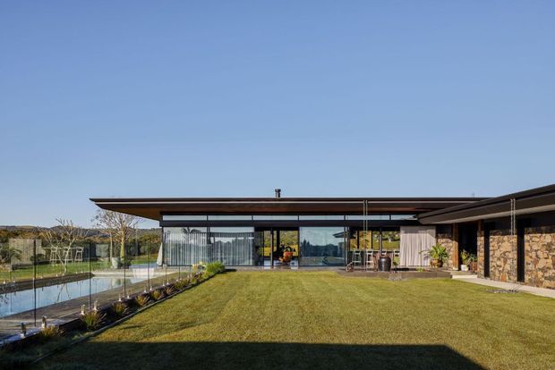 Wide exterior view of a low-slung modern residence arranged around a manicured lawn and reflecting pool. The home features broad floor-to-ceiling glazing, thin projecting roof planes, dark metal framing, and a natural stone-clad wall, with landscaped grounds and a clear sky.