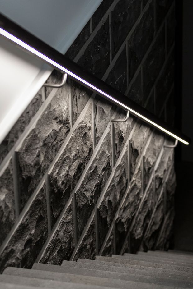 A dramatic close-up view down a narrow stairway. The stair wall is clad in irregular dark natural stone with light mortar joints, while a slim linear LED handrail/light runs diagonally along the wall. Grey textured stair treads and minimalist metal handrail brackets create a dark, contemporary architectural interior.