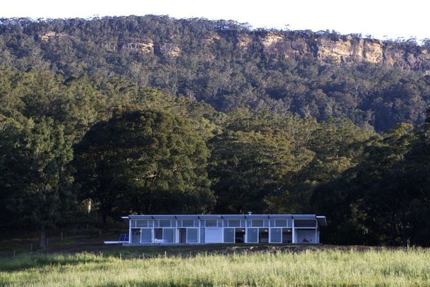 A low, elongated modern dwelling sits in a grassy clearing at the base of a densely forested escarpment. The building has extensive floor-to-ceiling glazing, a simple rectilinear form, and a shallow flat roof, creating a strong connection to the surrounding landscape.
