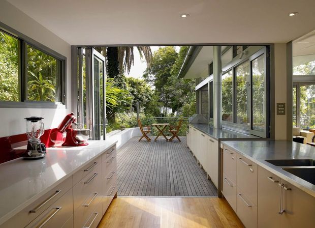 A bright contemporary kitchen opening directly onto a timber outdoor deck through wide glazed doors. The room has white built-in cabinetry and benchtops, dark-framed windows and doors, stainless-steel appliances, recessed ceiling lights, and a leafy garden setting beyond the deck.